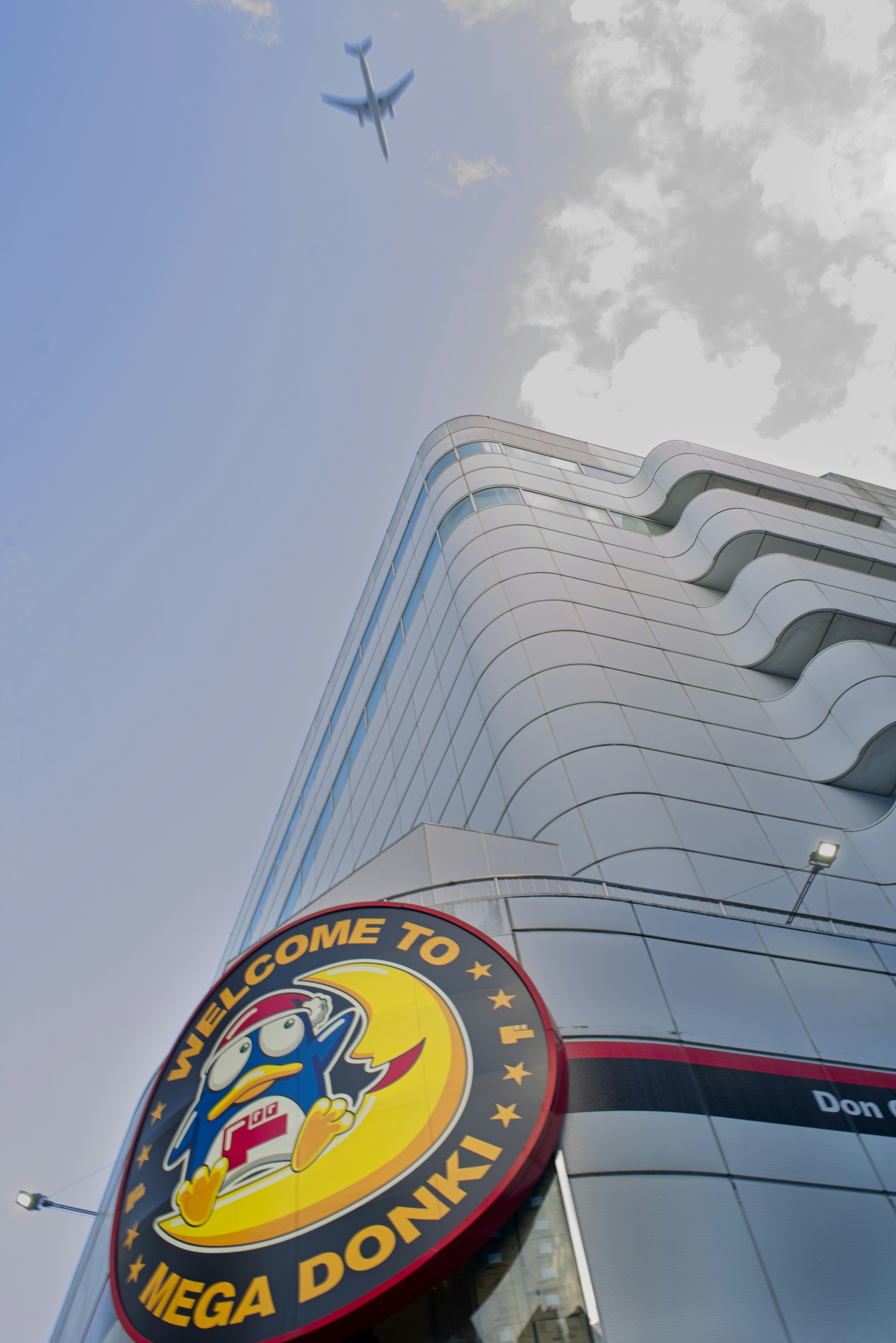 A quirky penguin mascot welcomes visitors to Mega Donki beneath a striking curved glass building with wave-like balconies rippling up its facade. Above, an airplane cuts across a sky split between clear blue and billowing white clouds, adding a surreal, dramatic contrast to the scene. Shot from a dramatic low angle, the composition emphasizes the building's sculptural architecture and the playful retail branding below, capturing an eye-catching corner of Shibuya on a hot July afternoon.